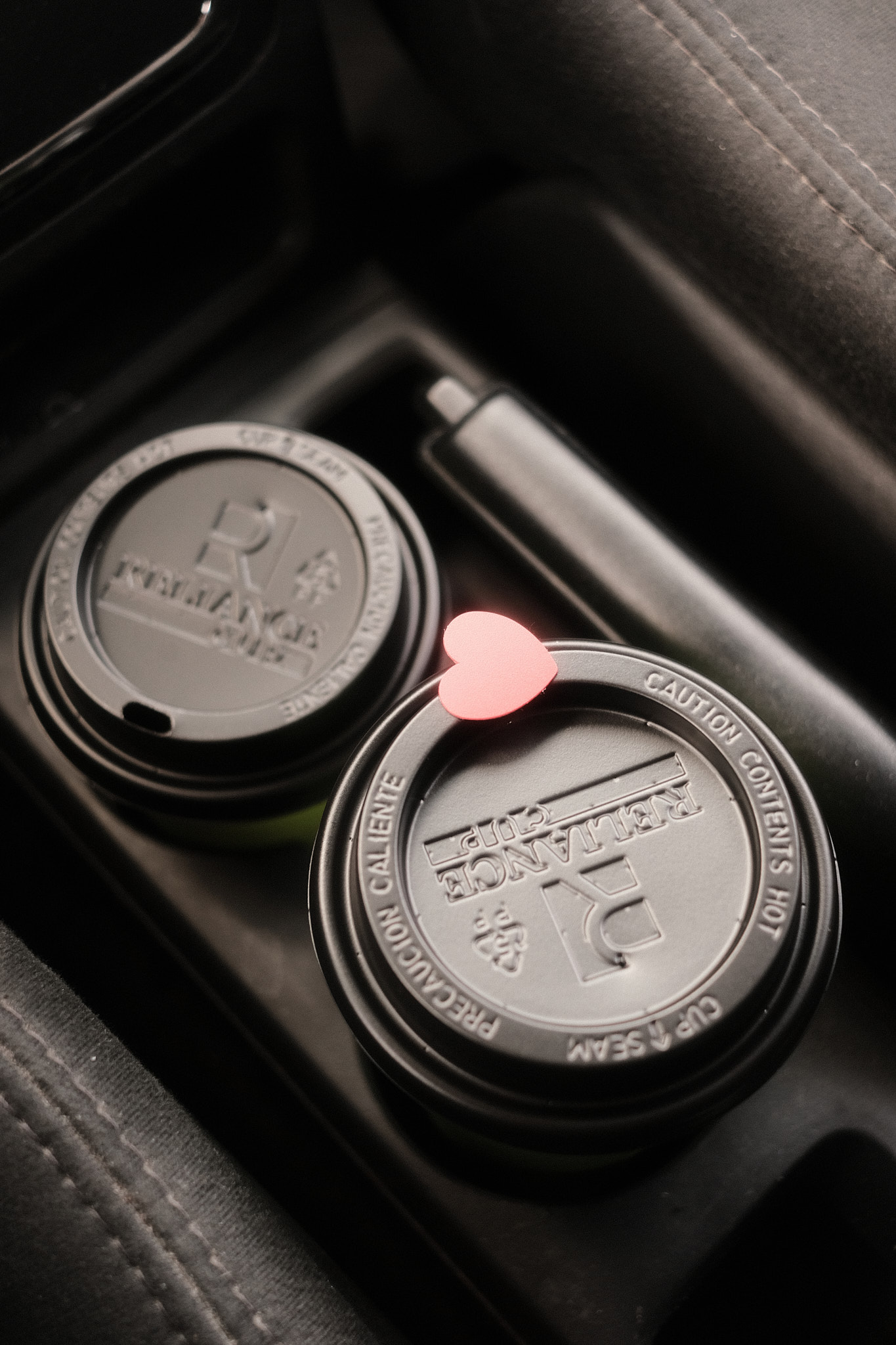 two cups of coffee shop coffee in a car holder of a car, the handbrake just beside it, and the lid of one with a heart sticker over the drink hole of the lid