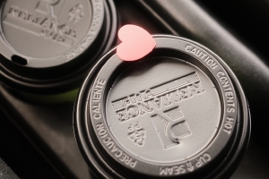 two cups of coffee shop coffee in a car holder of a car, the handbrake just beside it, and the lid of one with a heart sticker over the drink hole of the lid