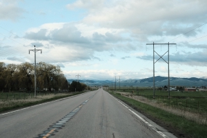 Driving down a two-lane highway at midmorning, flanked by power poles on each side, the sky covered lightly in clouds, and the mountains off in the distance.