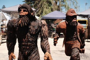 two wood carvings of Sasquatch, one without any clothes on and another with a cap, shorts, and a button up shirt that’s unbuttoned and opened