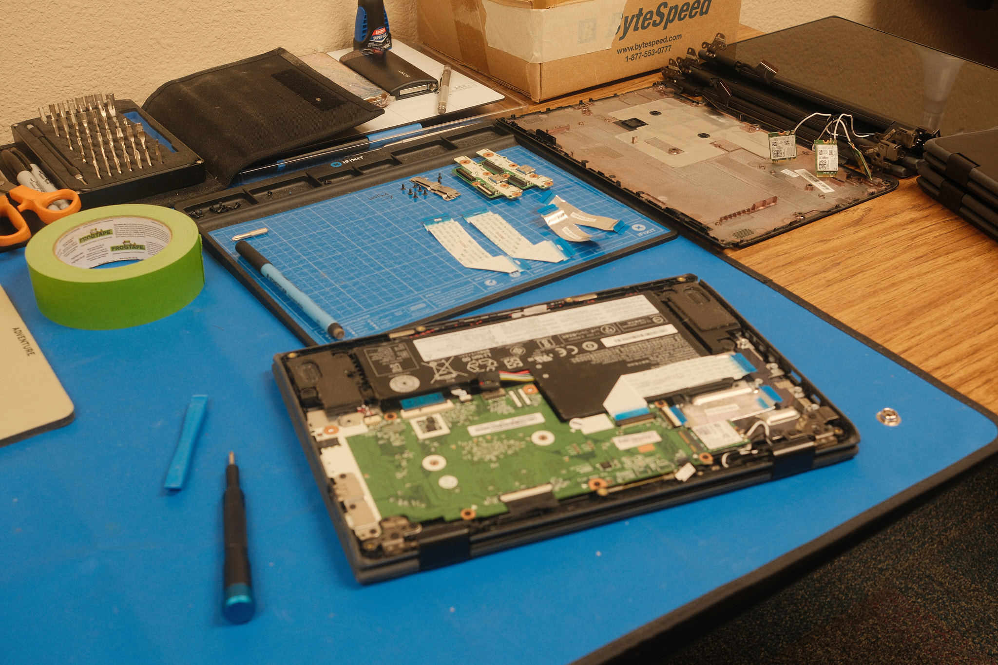 A work table with various tools and Chromebook parts on it. A Chromebook lies on it without its bottom cover, its battery and motherboard exposed.