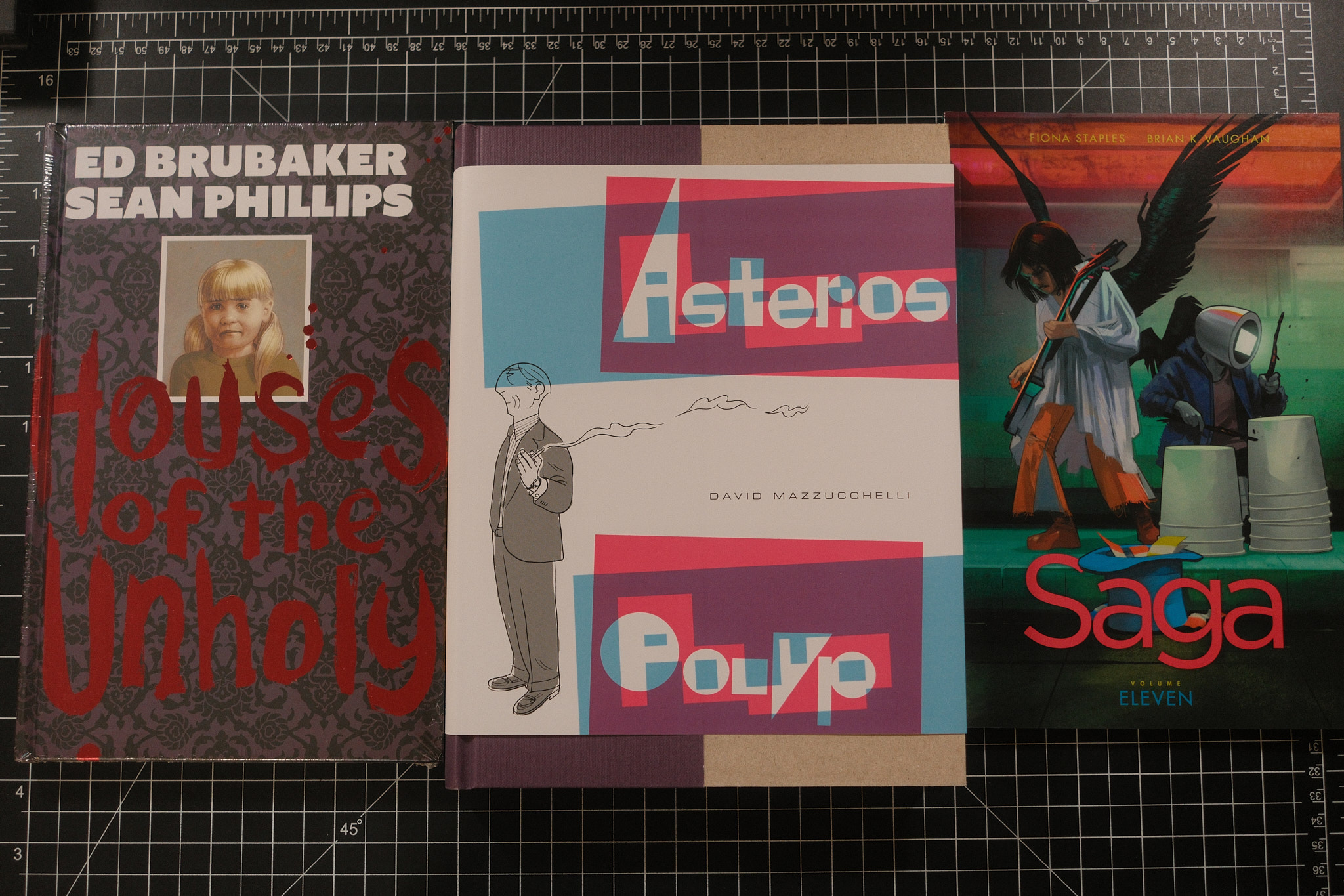 Three comic books sitting on a desktop. The three books are Houses of the Unholy by Ed Brubaker, Jacob Phillips, and Sean Phillips, Asterios Polyp by David Mazzucchelli, and Saga Volume 11 by Brian K. Vaughan and Fiona Staples