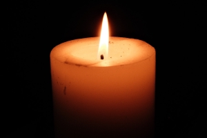 A single white pillar candle burning against a completely black background, its flame casting a warm light